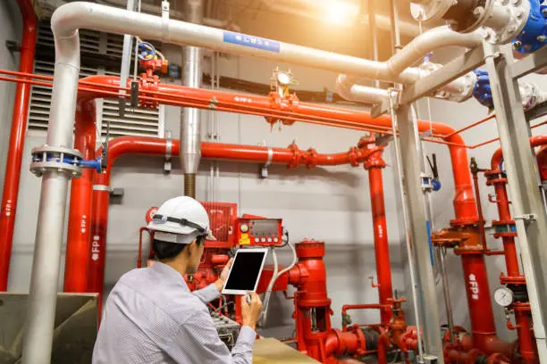 Person wearing a white safety helmet and light-colored shirt inspecting a red fire protection piping system in an indoor utility room. They hold a tablet device, likely used for recording data or following a maintenance checklist. The labeled pipes, valves, gauges, and control panels suggest a fire suppression or water distribution system.