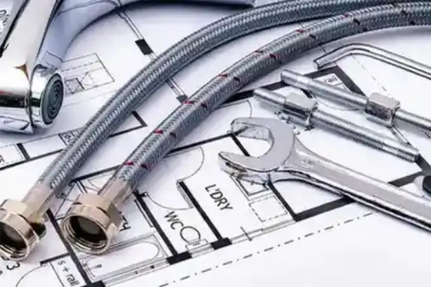 Plumbing tools and components—including braided metal hoses, a chrome faucet, wrench, and fasteners—arranged on architectural blueprints labeled with 'WC' and 'L'DRY'. The image highlights the planning and installation phase of MEP systems, specifically plumbing layout and fixture placement in a residential or commercial project