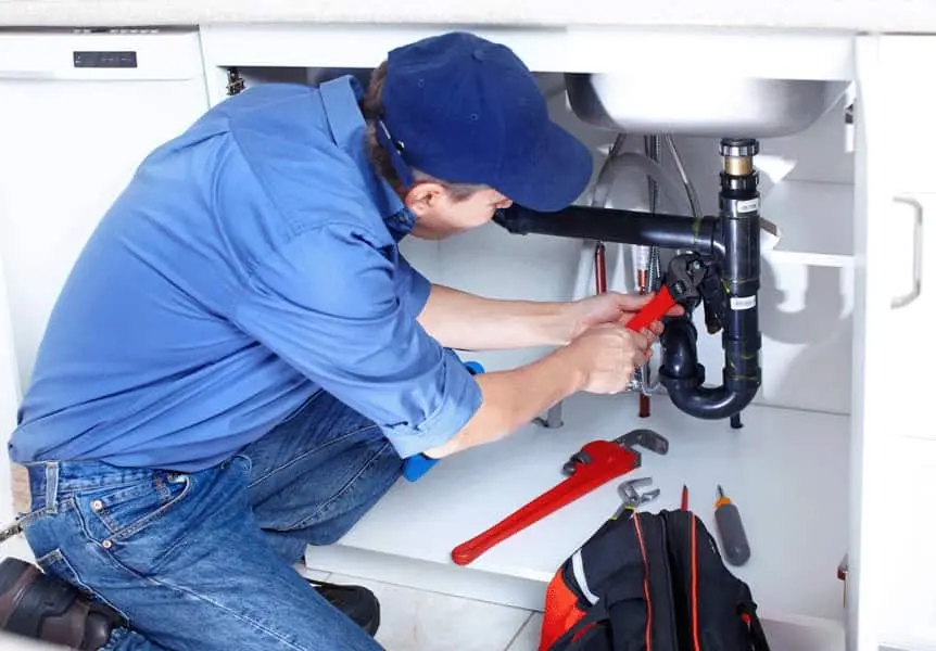 Plumbing service inqatar-markline trading & solutions Person in blue shirt and cap using a red pipe wrench to repair black PVC plumbing under a kitchen sink, with tools and a tool bag on the cabinet floor