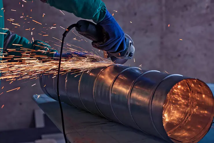 Person wearing protective gloves and a green sleeve using an angle grinder to cut a galvanized steel duct on a workbench. Bright sparks fly from the point of contact, highlighting active metalwork in an industrial workshop setting