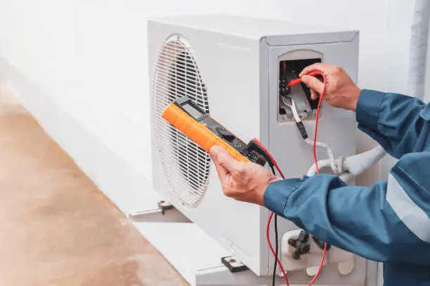 Technician in a blue uniform using a digital multimeter to test electrical components of an outdoor air conditioning unit mounted on a concrete surface beside a white wall