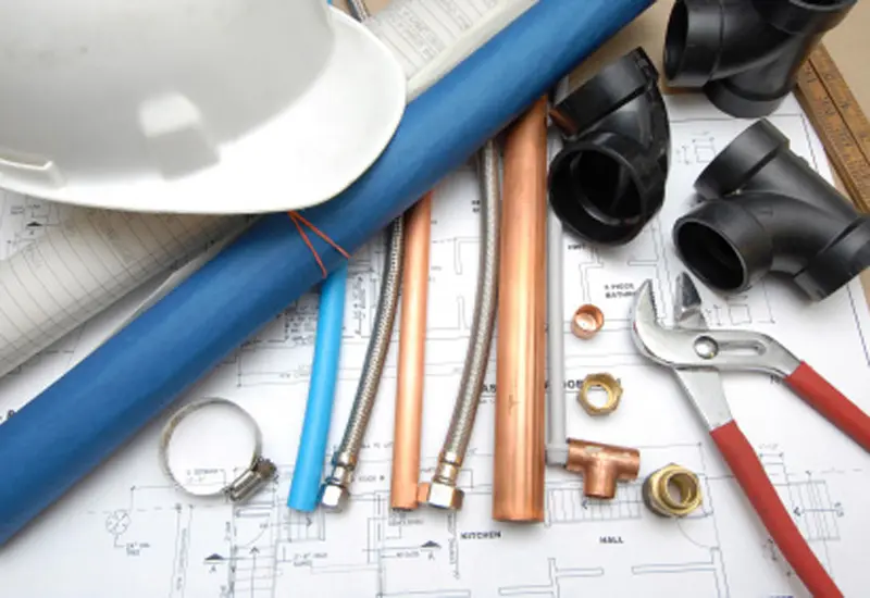 Plumbing tools and materials arranged on architectural blueprints, including pipes, fittings, a wrench, and a construction helmet