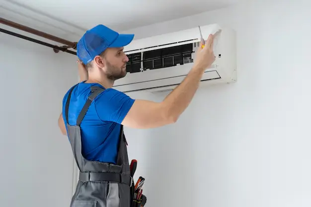 Markline -Air Conditioning companies in qatar A male air conditioning technician, wearing a blue cap and work overalls, is shown in a close-up shot as he services a white, wall-mounted AC unit. He is using a screwdriver to adjust the internal components of the unit. The image is a stock photo.