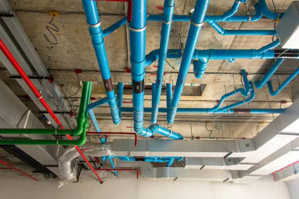 building piping services-markline trading & contracting Ceiling view of an industrial building under construction, showing a complex network of color-coded HVAC ductwork and piping. Blue, green, and red pipes indicate different utilities, while insulated metal ducts support air distribution. Exposed concrete and structural elements highlight the unfinished state of the site