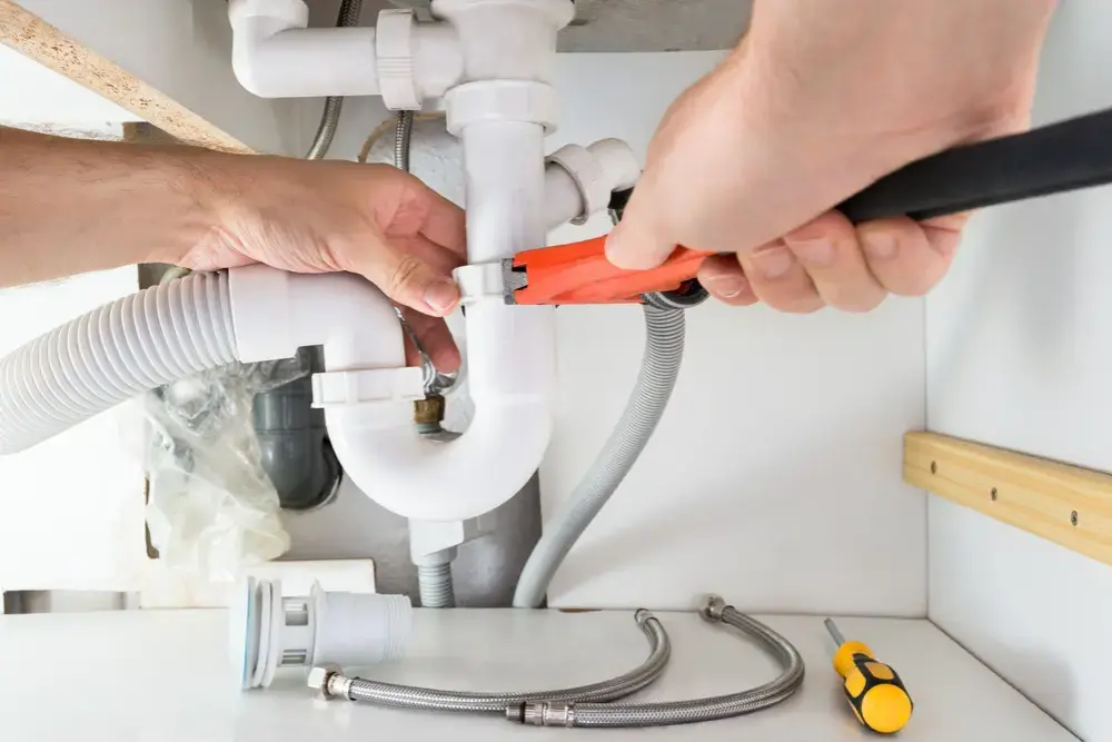 emergency plumbing services-markline trading & contracting Close-up of a person using a red-handled wrench to adjust white PVC pipes under a sink, with tools and plumbing parts scattered on the cabinet floor