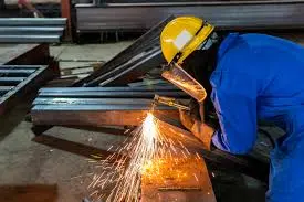 Welder in yellow hard hat and blue coverall creating sparks while working on metal in an industrial setting.