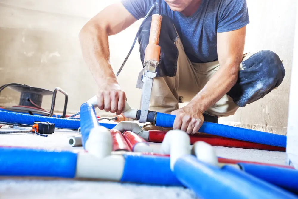 pipe installation-markline trading & contracting Person kneeling on a construction site floor, using a specialized tool to connect blue and red plastic plumbing pipes, indicating cold and hot water lines. Surrounding them are white pipe fittings, a measuring tape, and other plumbing components, suggesting active installation work in a building under renovation