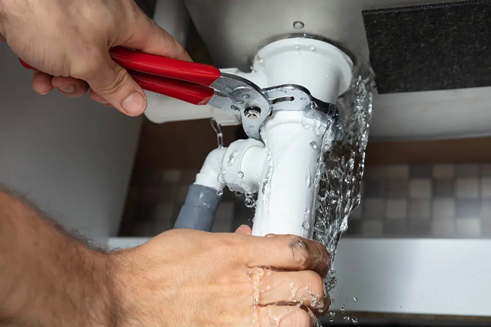 plumbing services in qatar-markline trading Close-up of a person using a red-handled wrench to fix a leaking white PVC pipe under a sink, with water spraying from the joint.