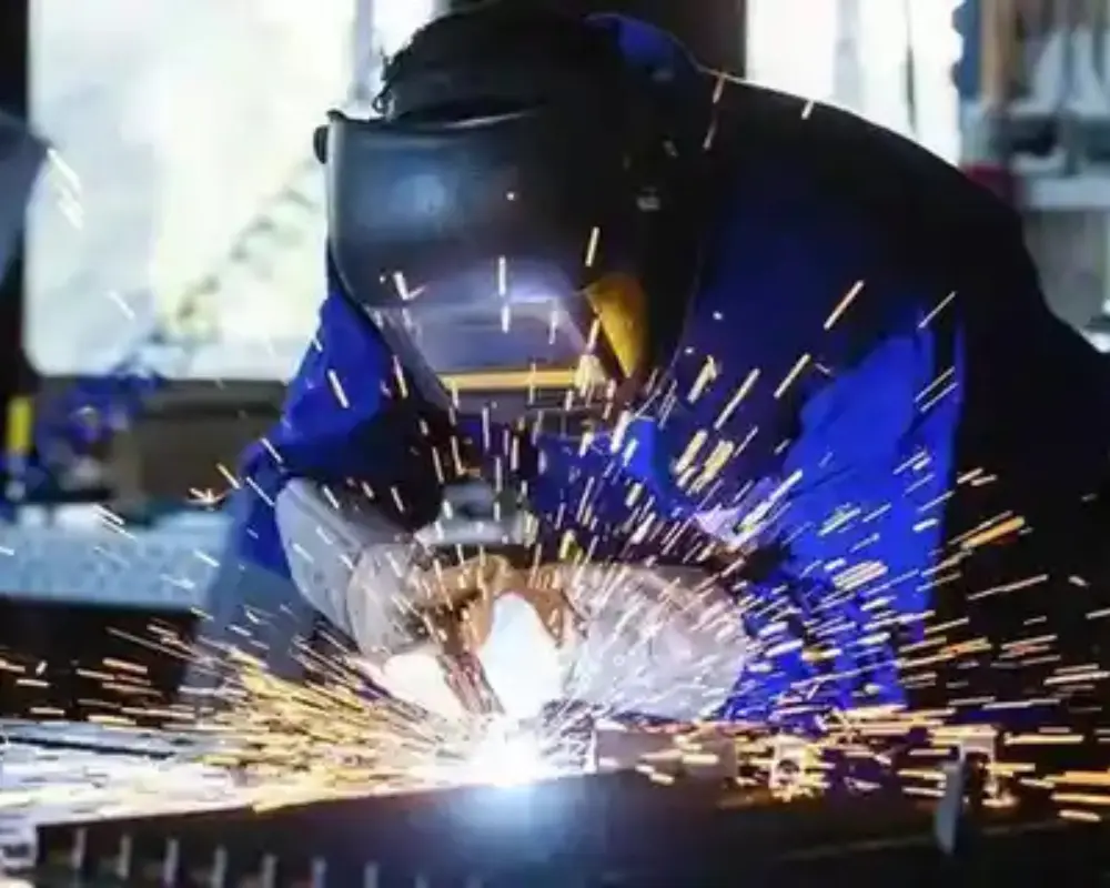 Person wearing a welding helmet and protective suit actively welding metal, with bright sparks flying in an industrial workshop setting.<br />
