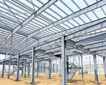 Steel framework of a large building under construction, featuring vertical columns, horizontal beams, roof trusses, and an installed staircase on a sandy ground.