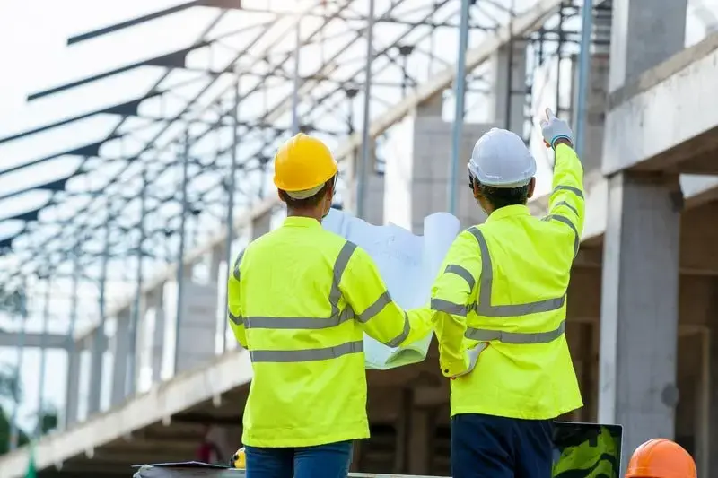 MEP companies in Qatar- Markline Trading Contracting (1) Two construction professionals, viewed from the back, are on a construction site. Both are wearing fluorescent yellow safety vests and hard hats (one yellow, one white). One person holds a blueprint or building plan and the other is pointing up and out toward the building's structure or skeleton in the background, suggesting they are discussing the plans.